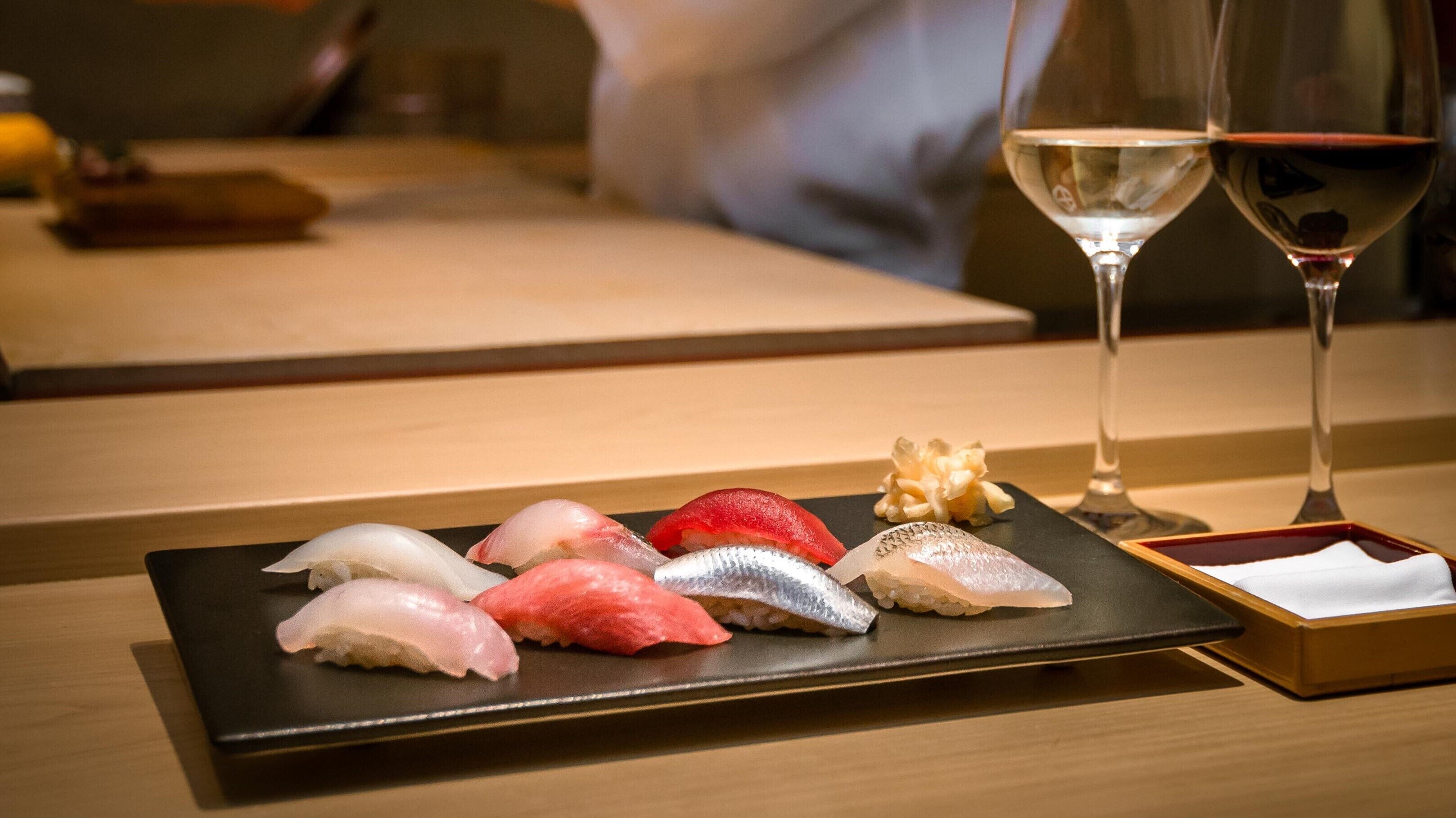 A sushi platter with various types of fish on a black rectangular plate, accompanied by a glass of red wine and a small wooden box with a white napkin.