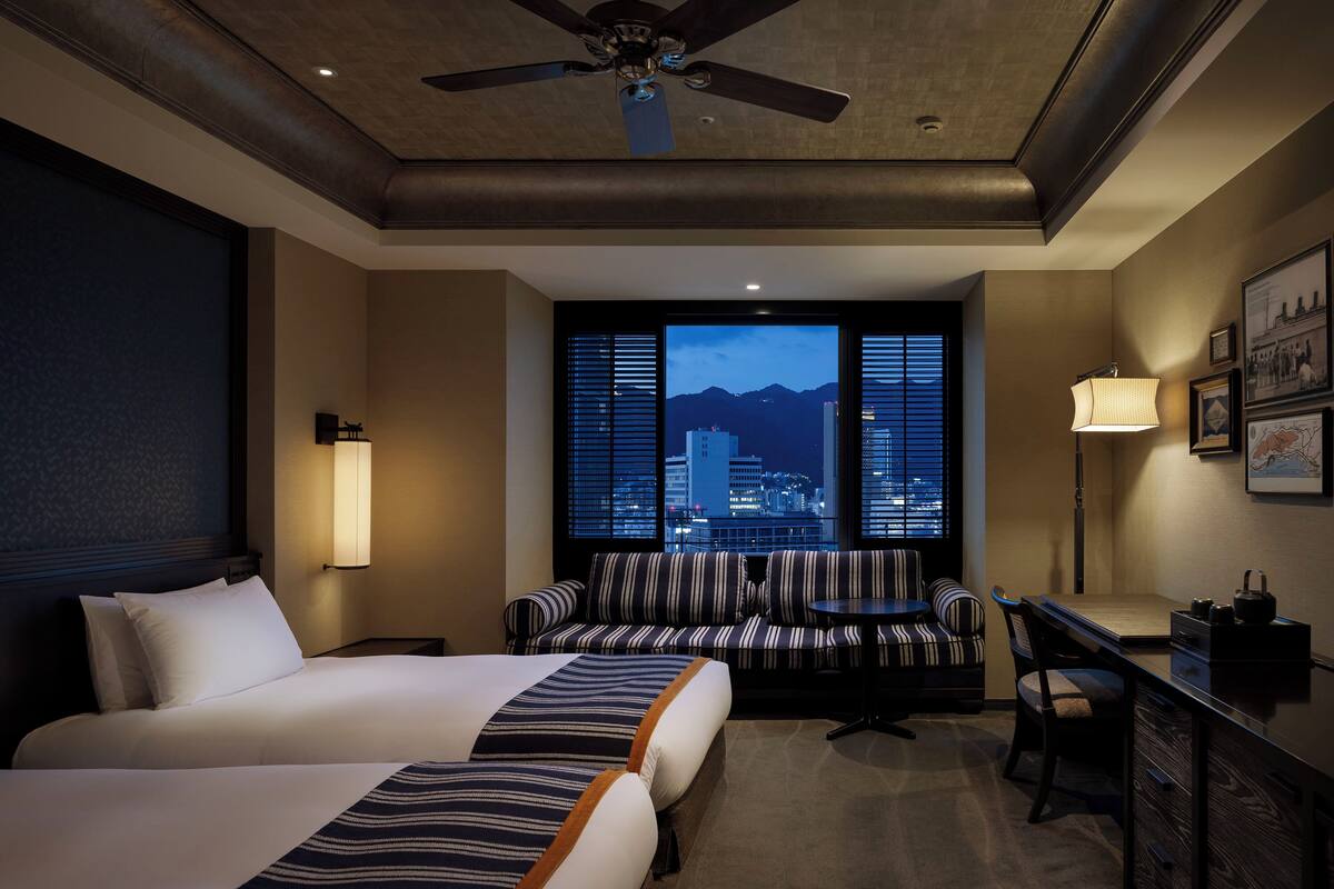 Habitación de hotel con cama, zona de estar con sofás a rayas, escritorio y vista a la ciudad de noche.