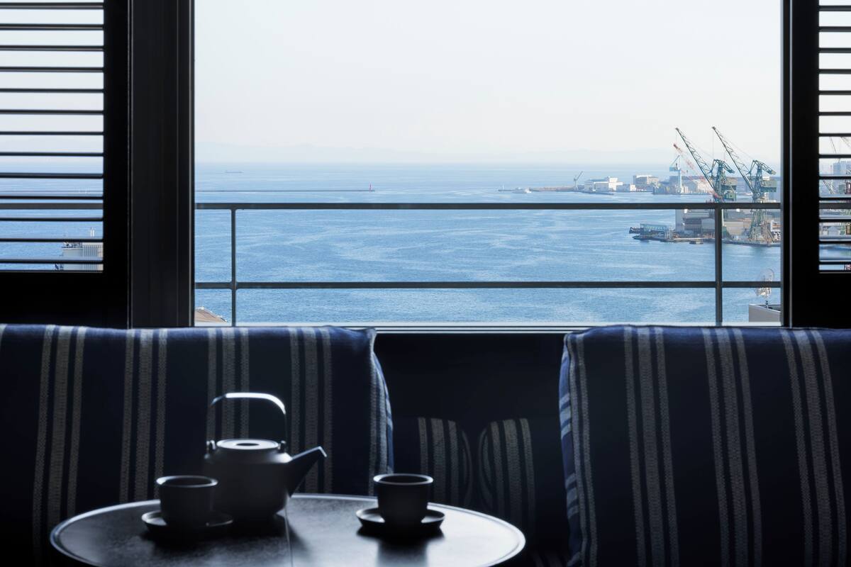 Vista desde un balcón con mesa y sillas, una tetera y tazas, con vista a un puerto con grúas y barcos.