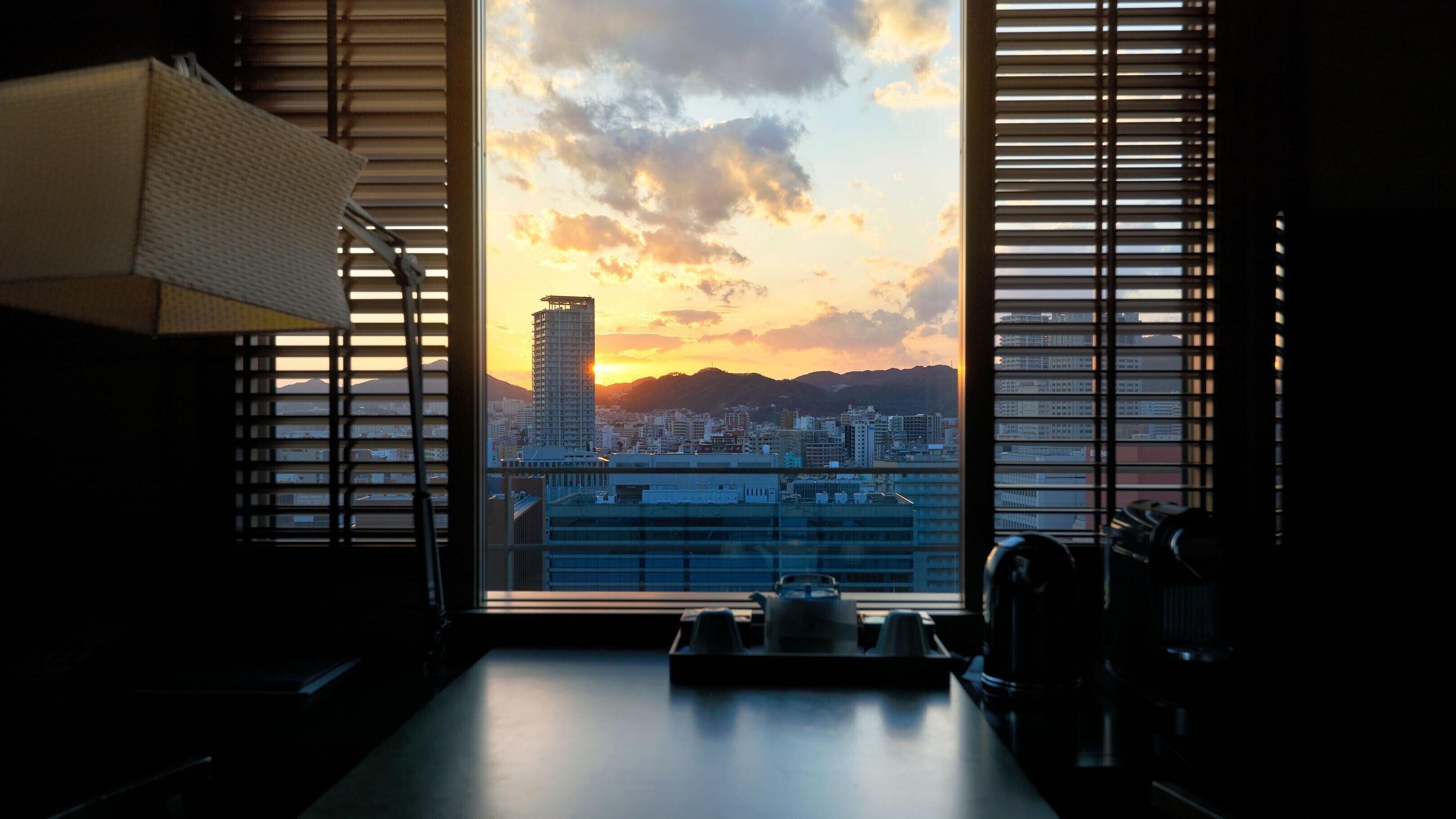 A room with a view of a cityscape at sunset through a window with blinds.