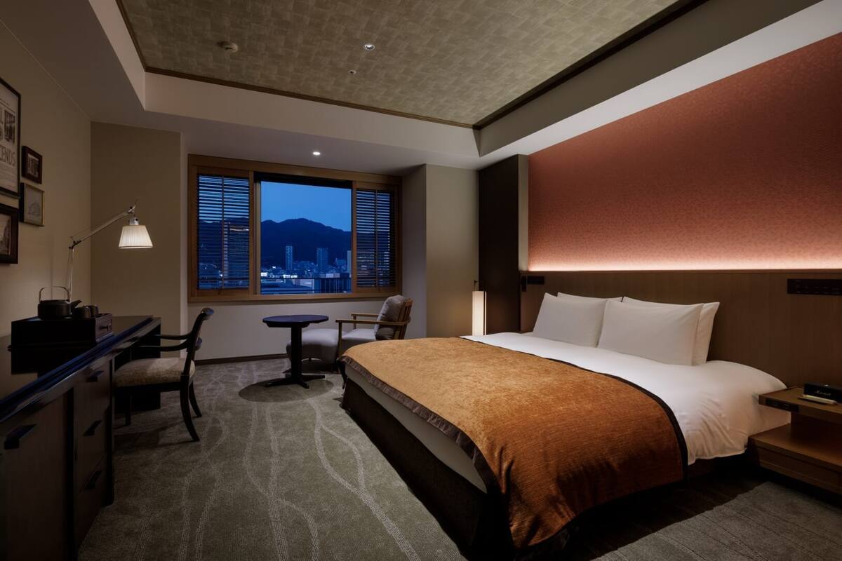 Habitación de hotel con cama, escritorio, silla y vista a un paisaje urbano a través de un ventanal amplio.