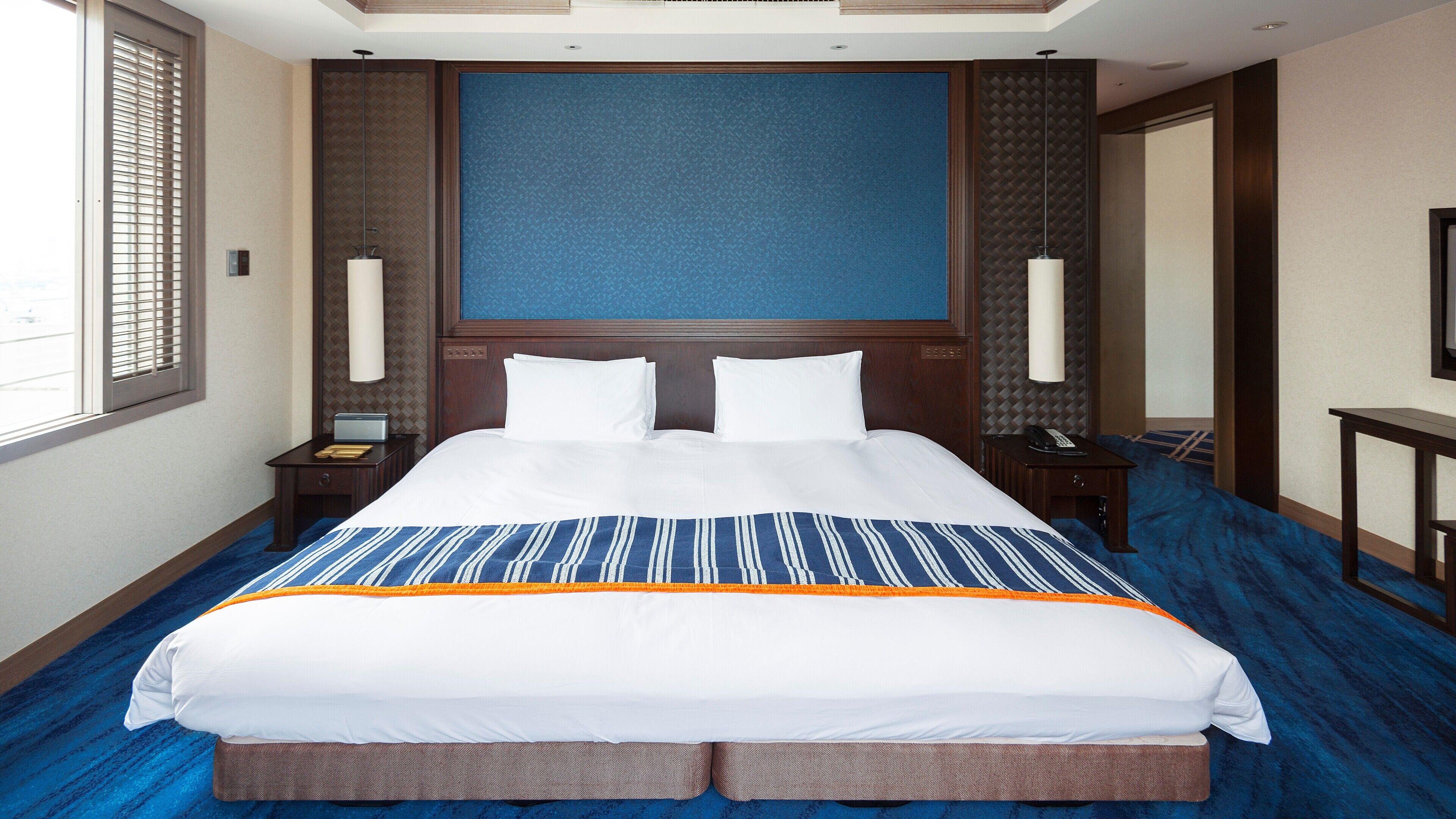 A hotel room with a large bed, a nightstand, a lamp, and a window with blinds.