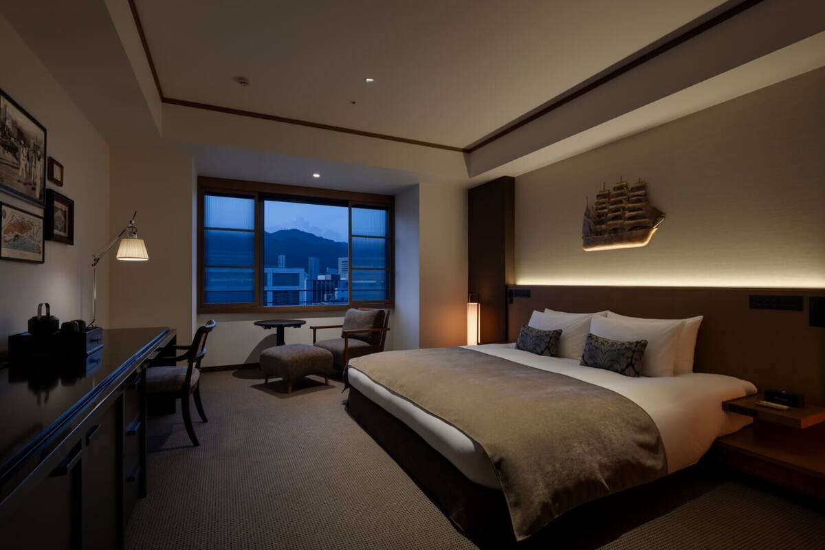 Habitación de hotel con una cama grande, un escritorio, una silla, una mesita y una ventana con vistas a la ciudad.