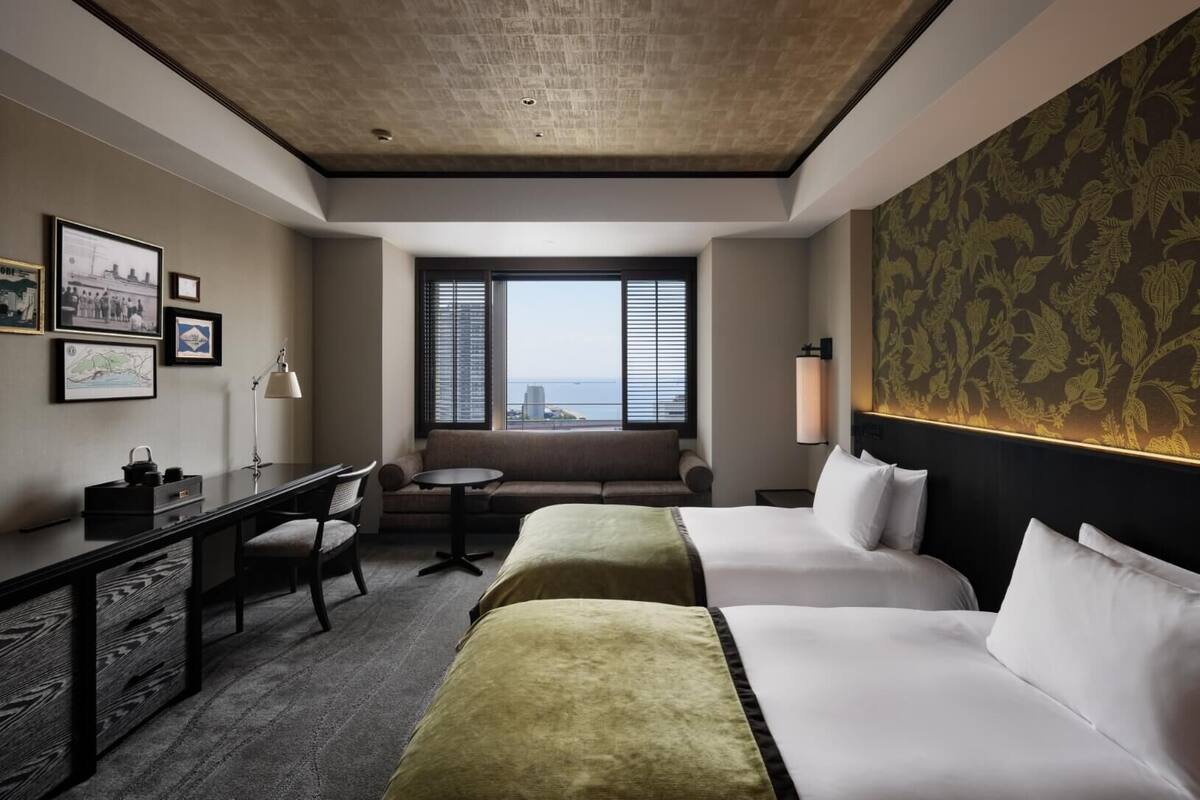 Habitación de hotel con dos camas, un escritorio, una silla, un sofá, una lámpara y una ventana con vistas a la ciudad.