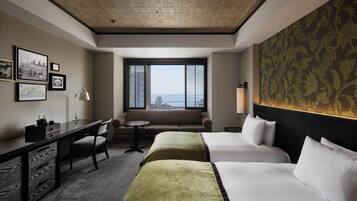 A hotel room with two beds, a desk, a chair, a sofa, a lamp, and a window with a city view.
