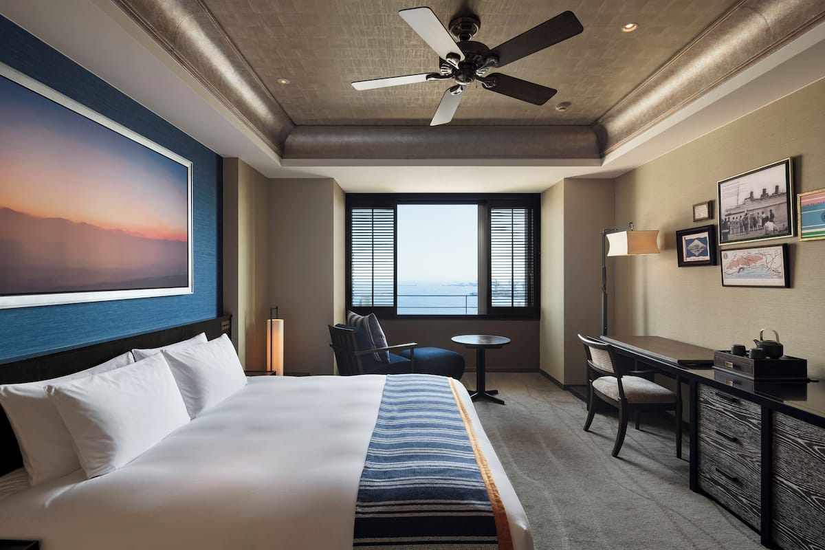 Una habitación de hotel con cama, escritorio, silla, ventilador de techo y una ventana grande.