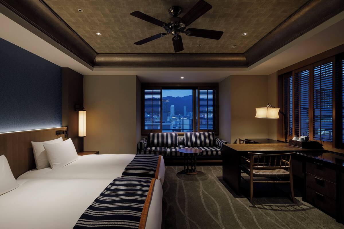 Habitación de hotel con cama, escritorio, sillón, sofá y vistas a la ciudad.
