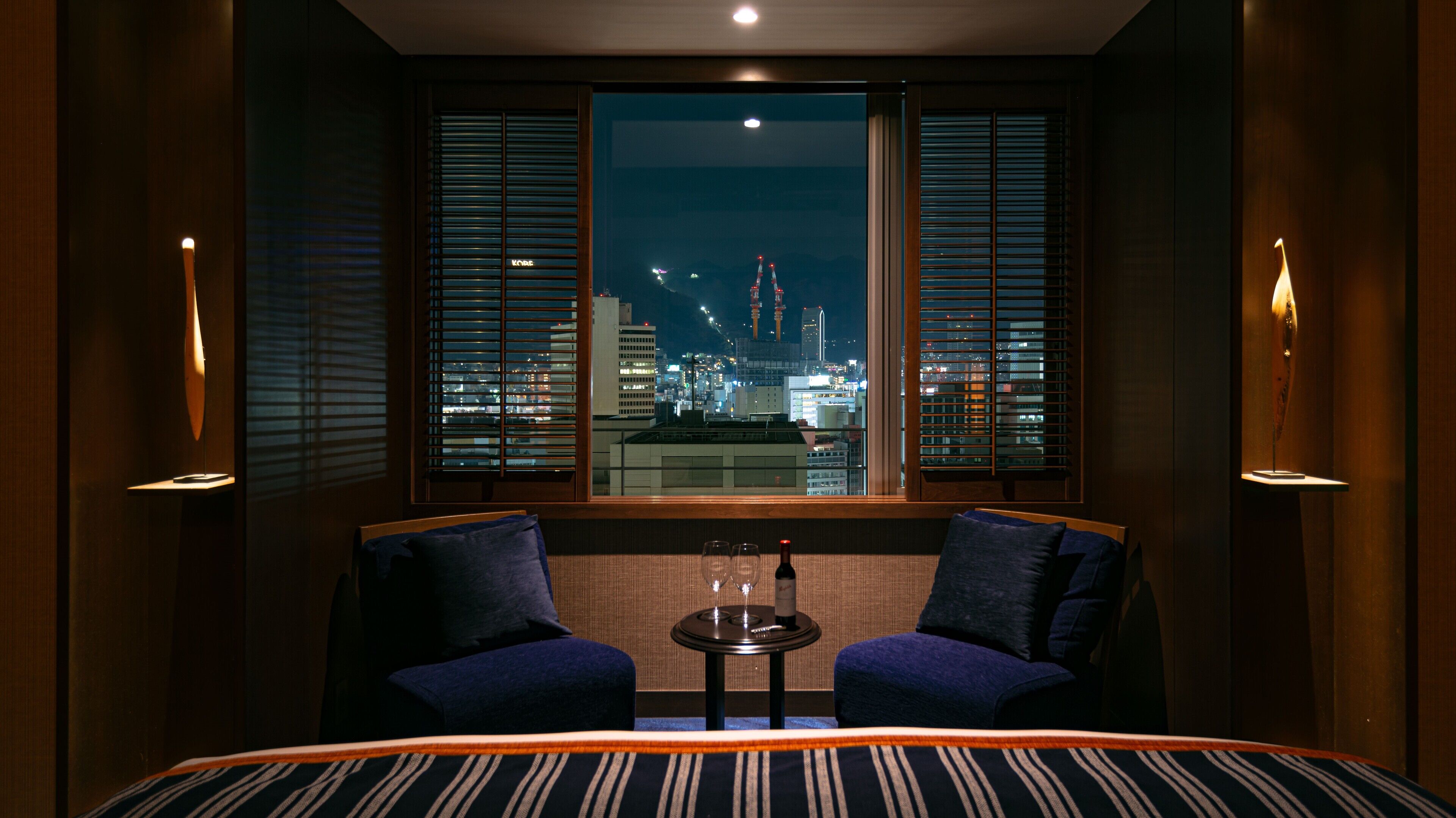 A hotel room with a large bed, two armchairs, a small table with a bottle and glasses, and a view of a cityscape at night.