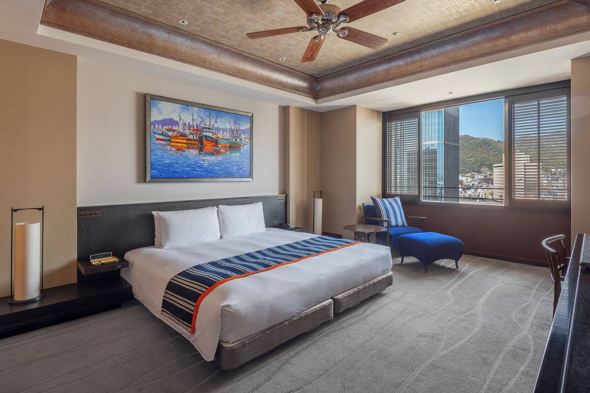 Habitación de hotel con una cama grande, un cuadro de barcos, vistas a la ciudad, un ventilador de techo y una ventana con persianas.
