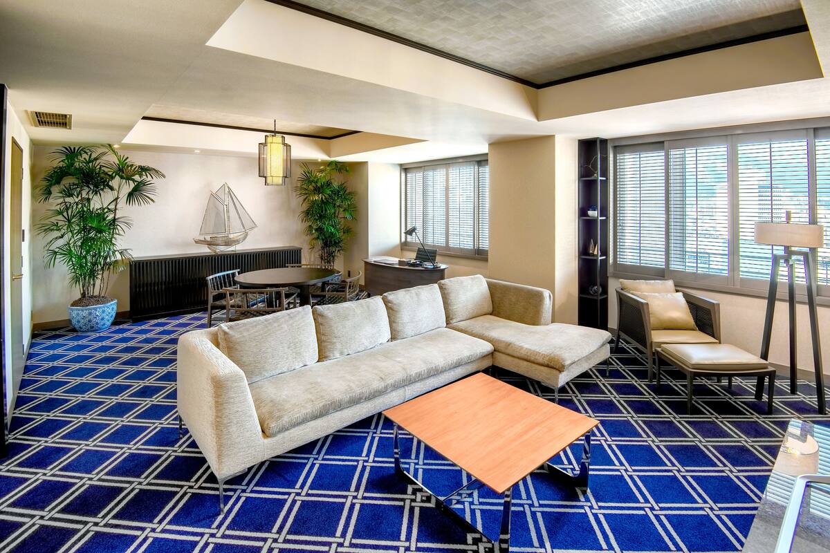 Una habitación de hotel moderna con un sofá modular, una pequeña mesa de madera, un rincón comedor y grandes ventanas con persianas.
