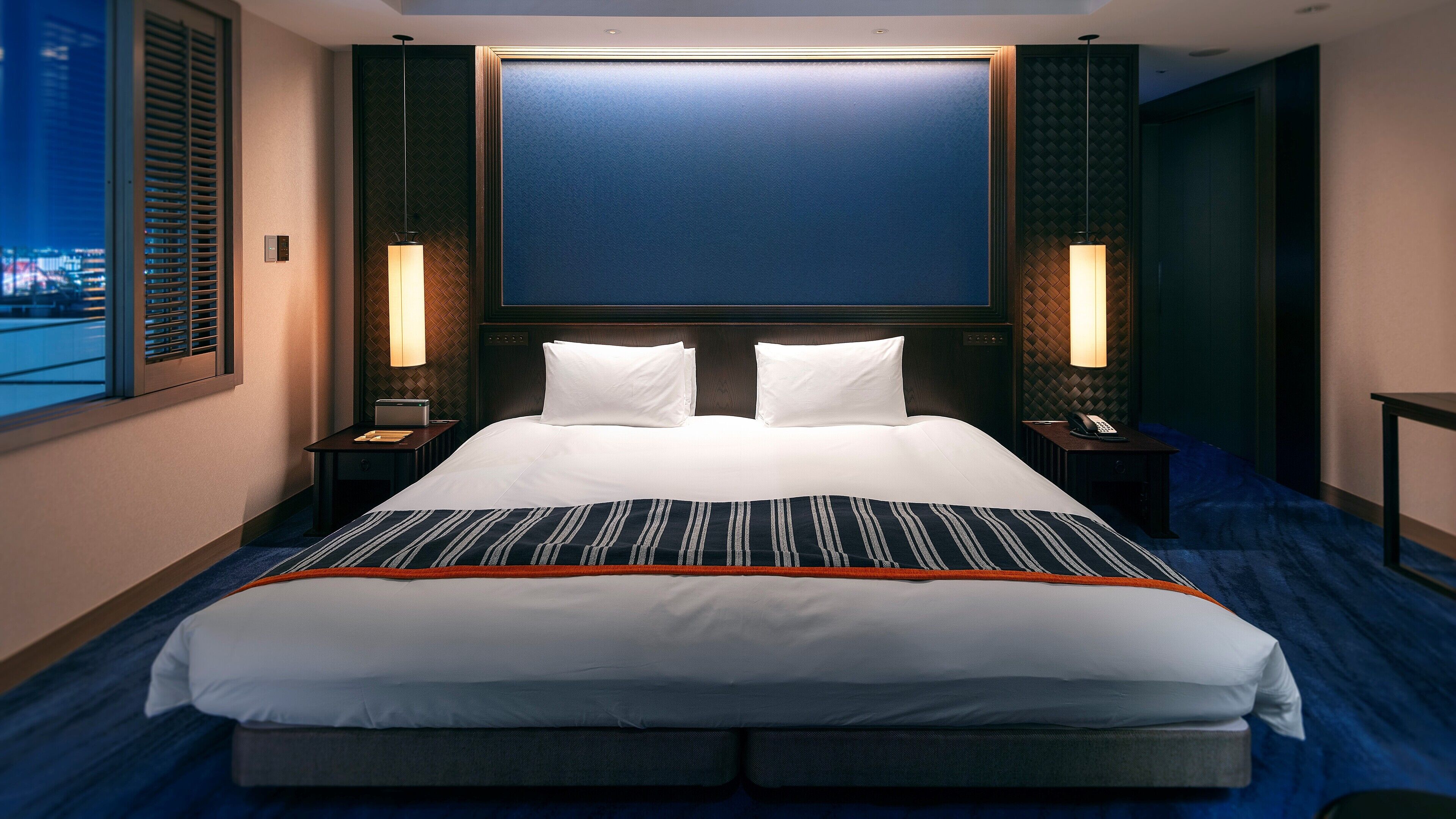 A hotel room with a large bed, two bedside tables, a nightstand, a wall-mounted TV, and a window with blinds.