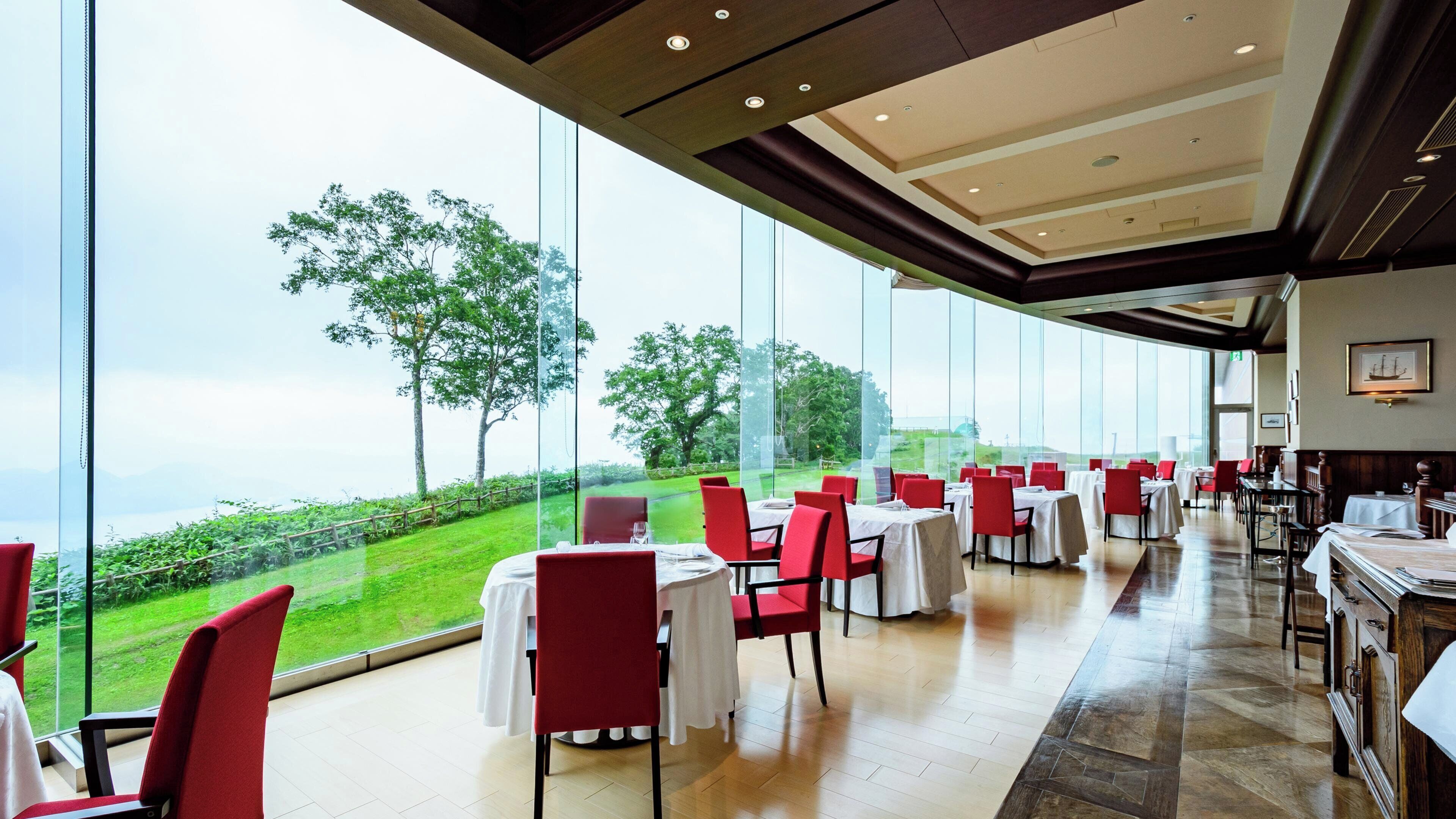 Un restaurante con amplios ventanales que ofrece vistas a un paisaje verde y árboles. Las mesas están preparadas con manteles blancos y sillas rojas.