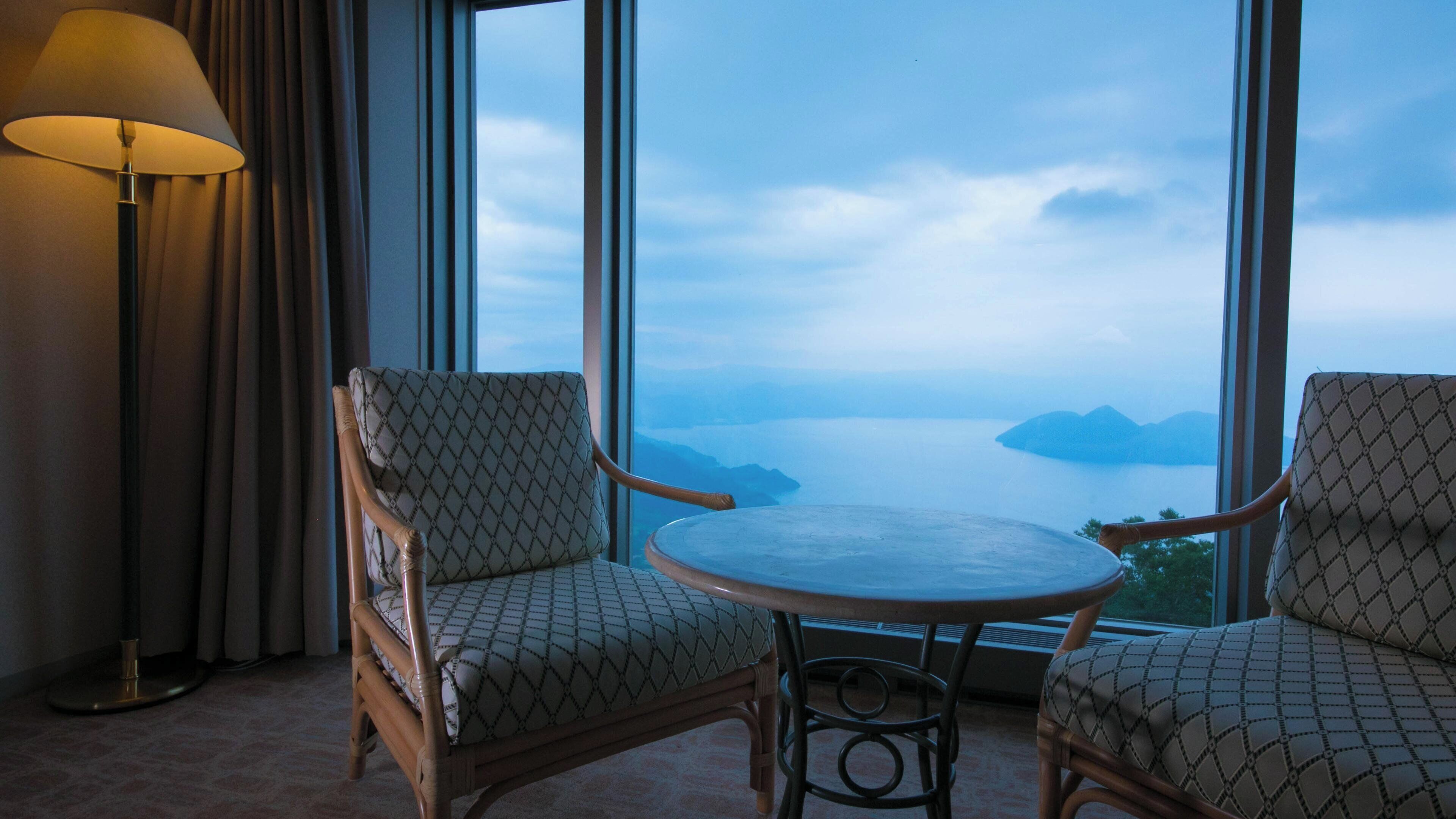 A room with two wicker chairs and a small round table, large windows with a view of a body of water and distant land.