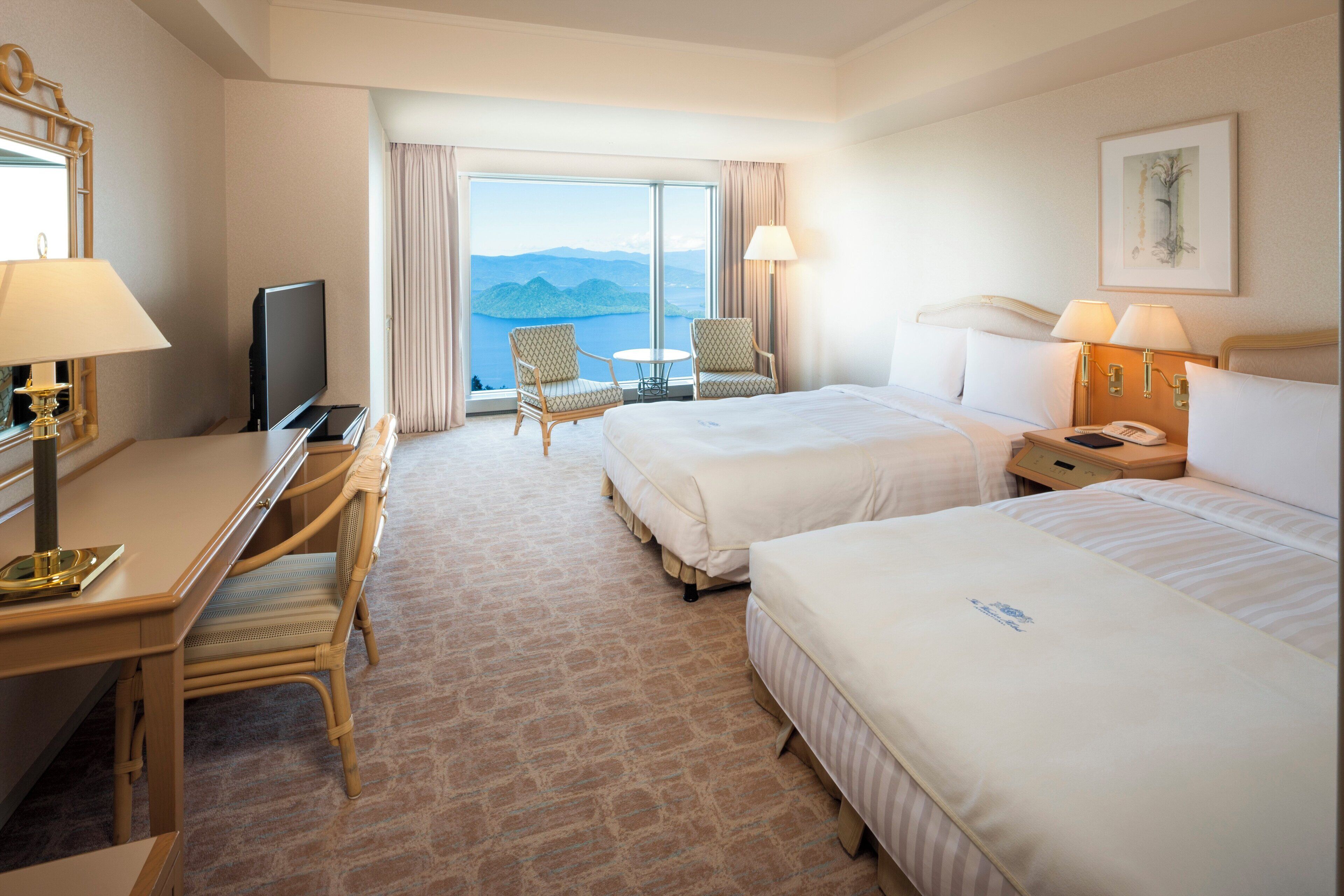 A hotel room with two beds, a desk with a TV, a chair, a lamp, and a view of the mountains.