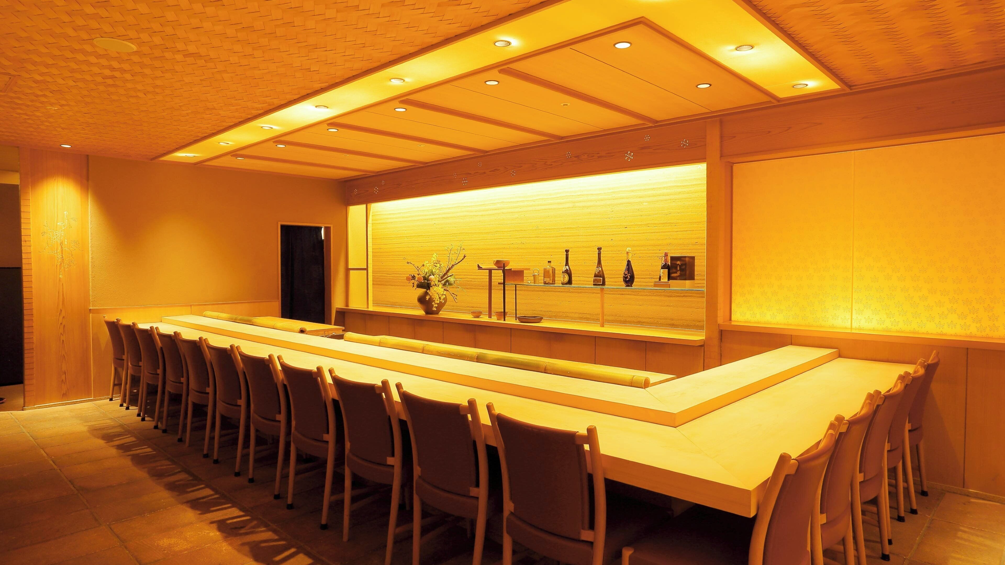 A long, rectangular bar area with a series of chairs around it, a wooden ceiling with recessed lighting, and a wall-mounted shelf with various bottles and a vase.