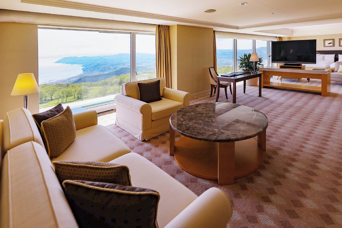 A spacious living room with a large window offering a scenic view, a comfortable sofa set, a coffee table, and a flat-screen TV.