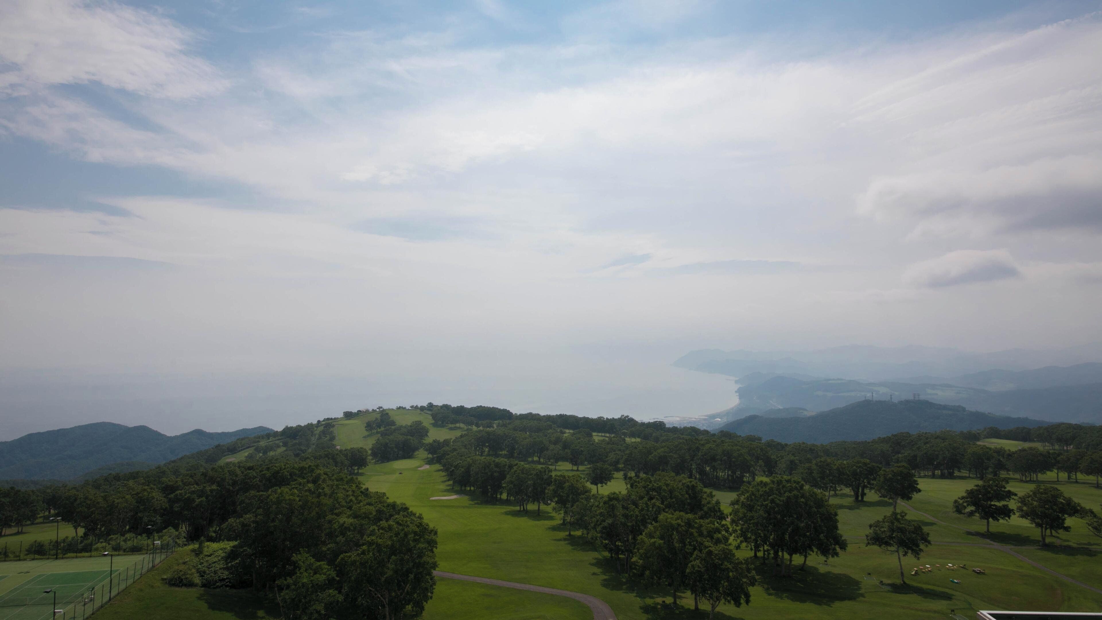 A golf course with a path leading through it, surrounded by trees and hills in the background.