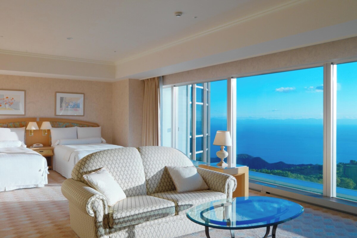 A hotel room with a king-sized bed, a sofa, a glass coffee table, and a large window offering a view of the ocean.