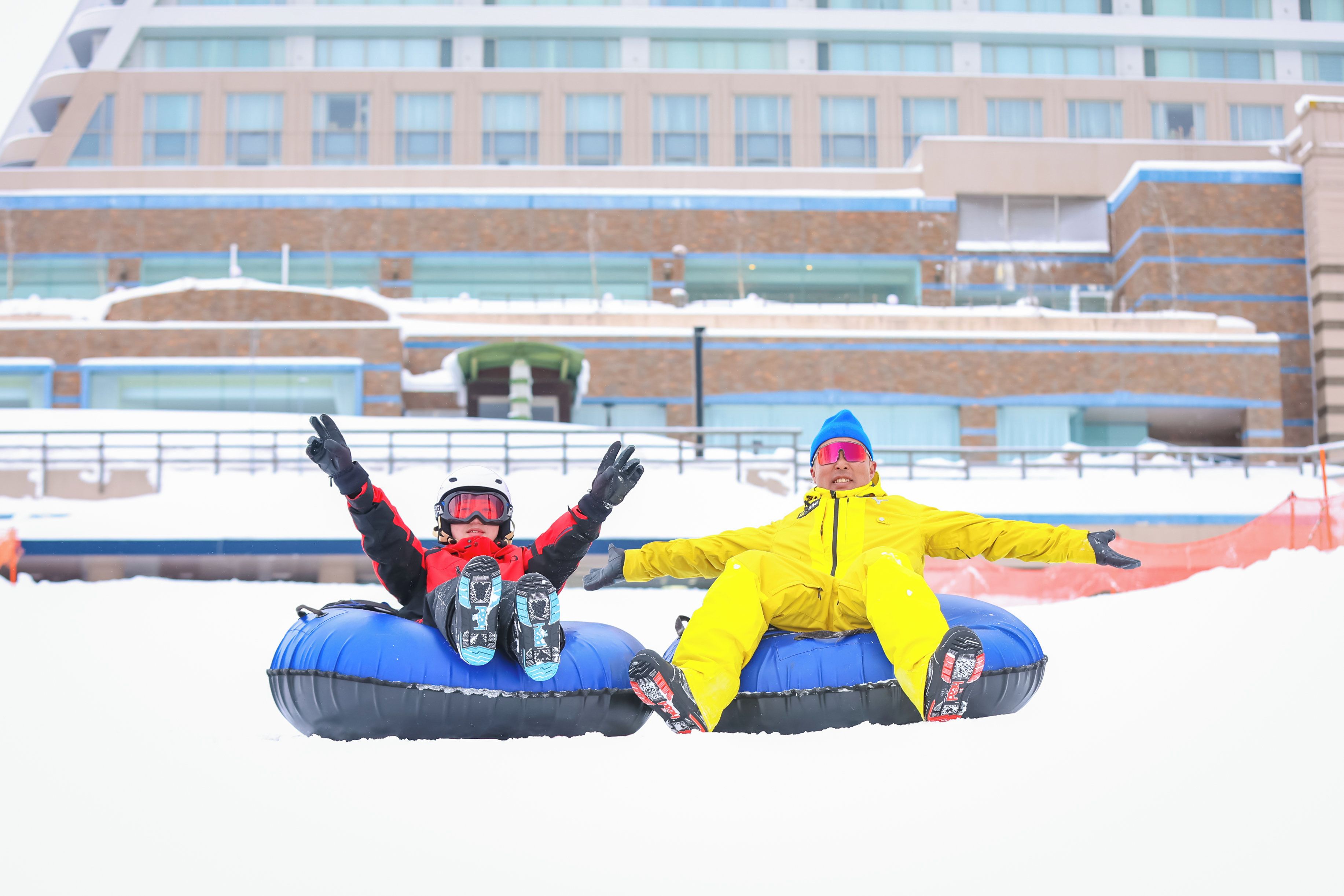 Two people on snow tubes in front of a building with a snow-covered ground.
