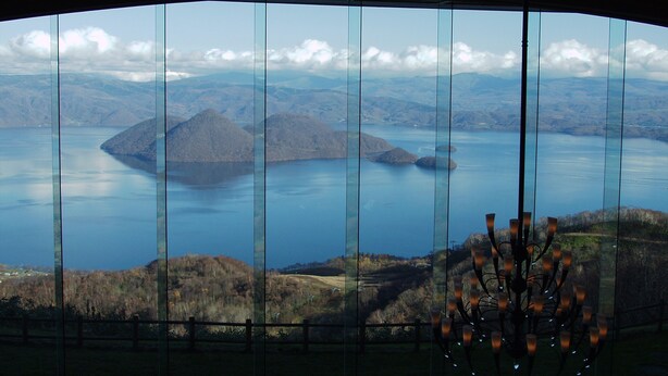 A panoramic view through a curved window, featuring a lake surrounded by hills and a distant mountain range. - The Windsor Hotel Toya, Vignette Collection by IHG (Toyako)
