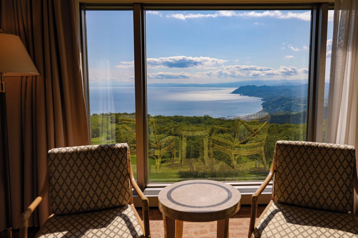 Um quarto com duas cadeiras estampadas e uma pequena mesa, uma janela grande com vista para a paisagem costeira e montanhas.