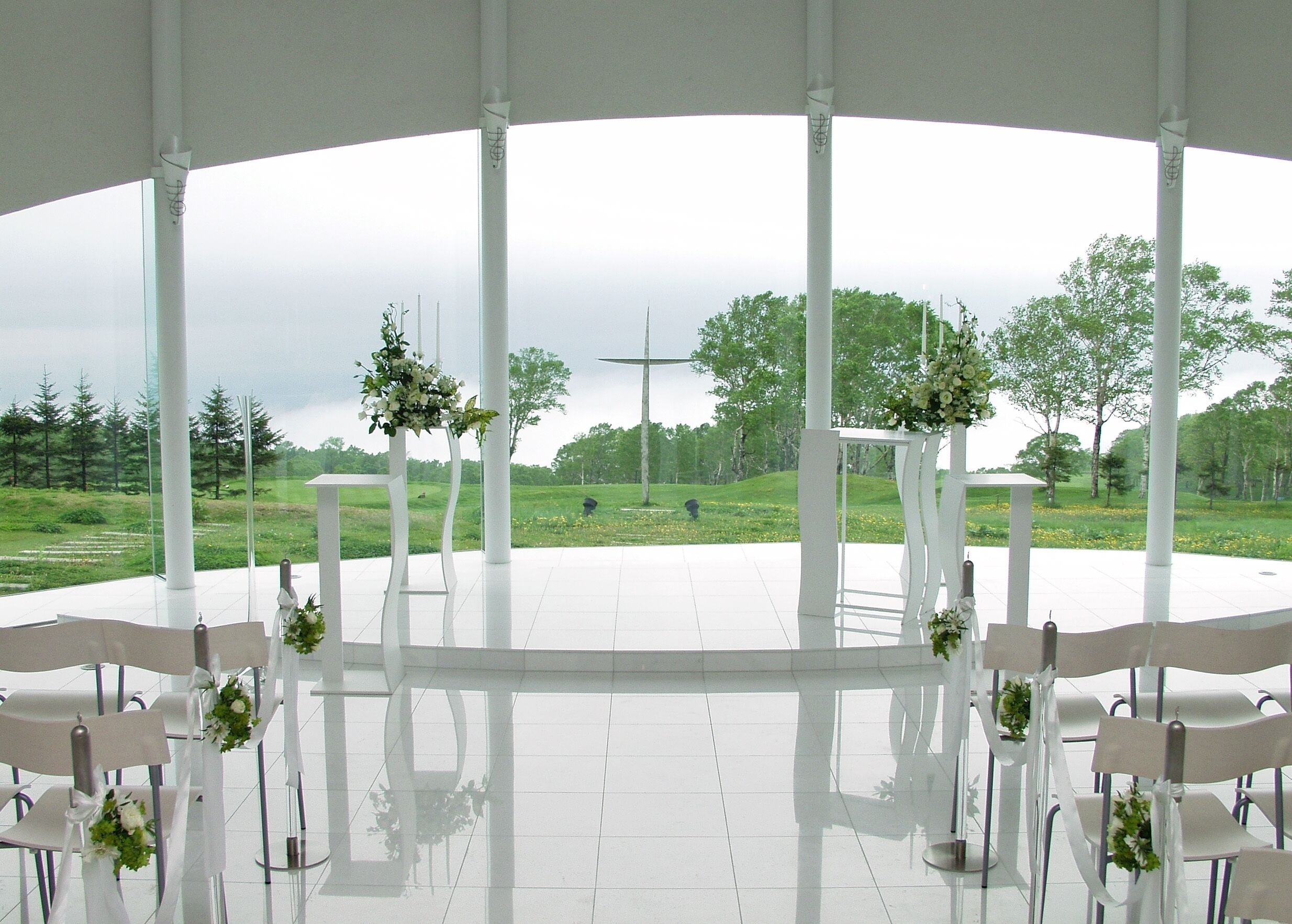 Un espacio al aire libre cubierto con sillas y mesas blancas, dispuesto para un evento, con vistas a un campo cubierto de césped y árboles.