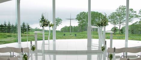 Un espacio al aire libre cubierto con sillas y mesas blancas, dispuesto para un evento, con vistas a un campo cubierto de césped y árboles.