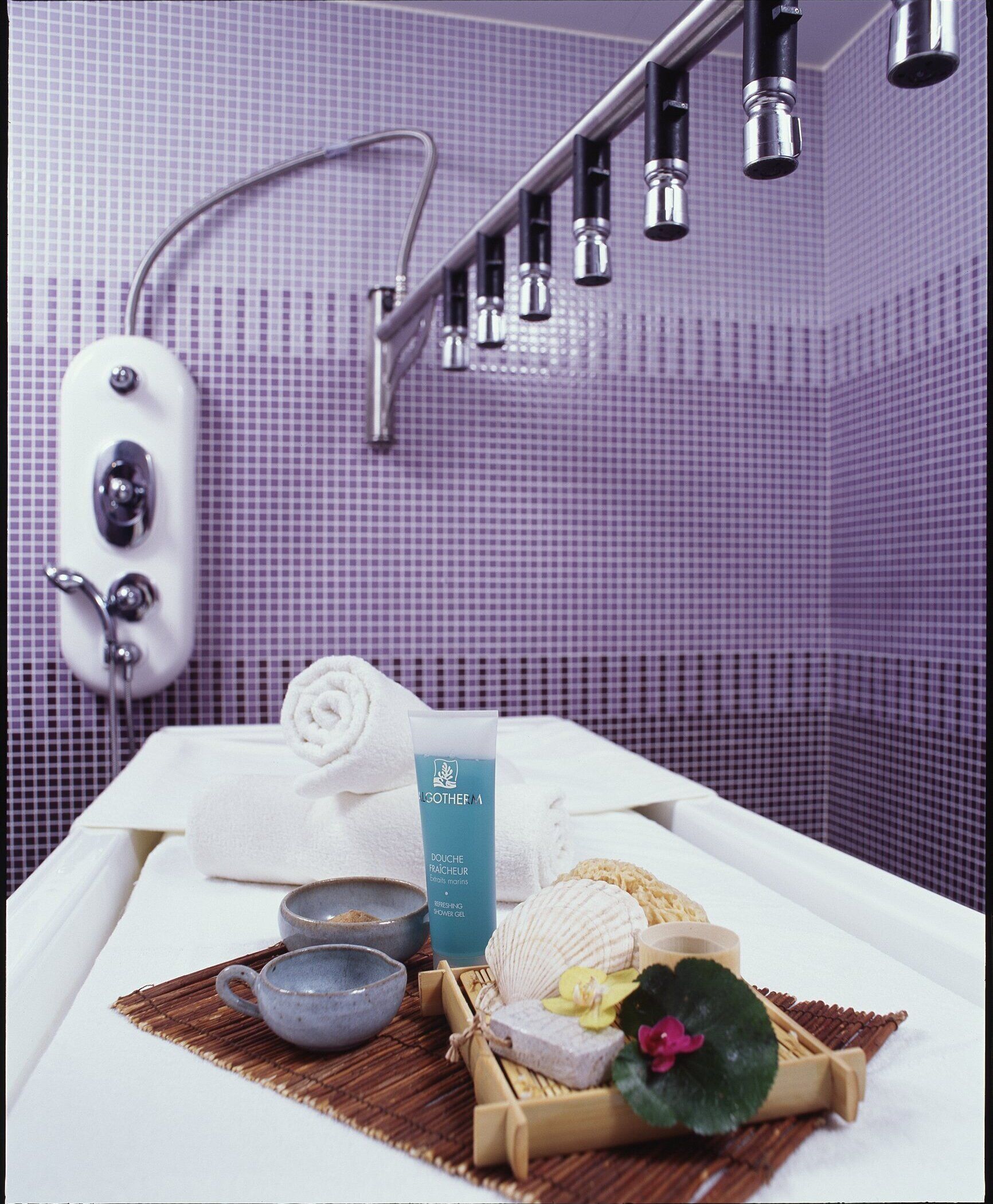 Een badkamer met een douche, een handdoek, een fles douchegel, en een dienblad met een beker, zeep en een bloem.