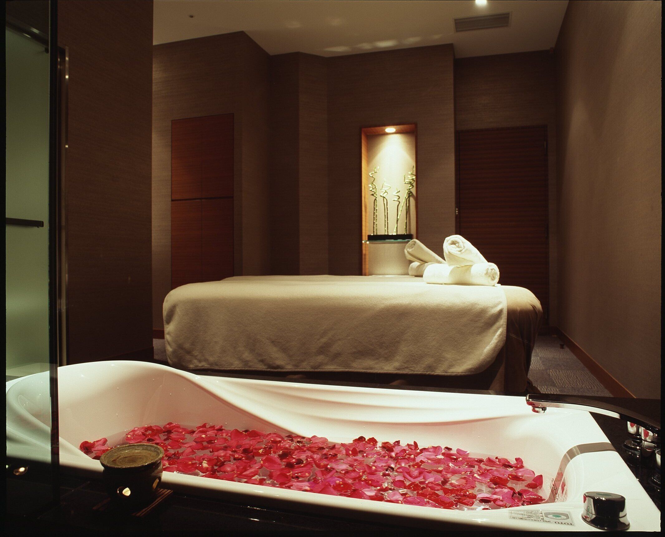 A hotel room with a bed, a bathtub filled with rose petals, and a wall-mounted light fixture.