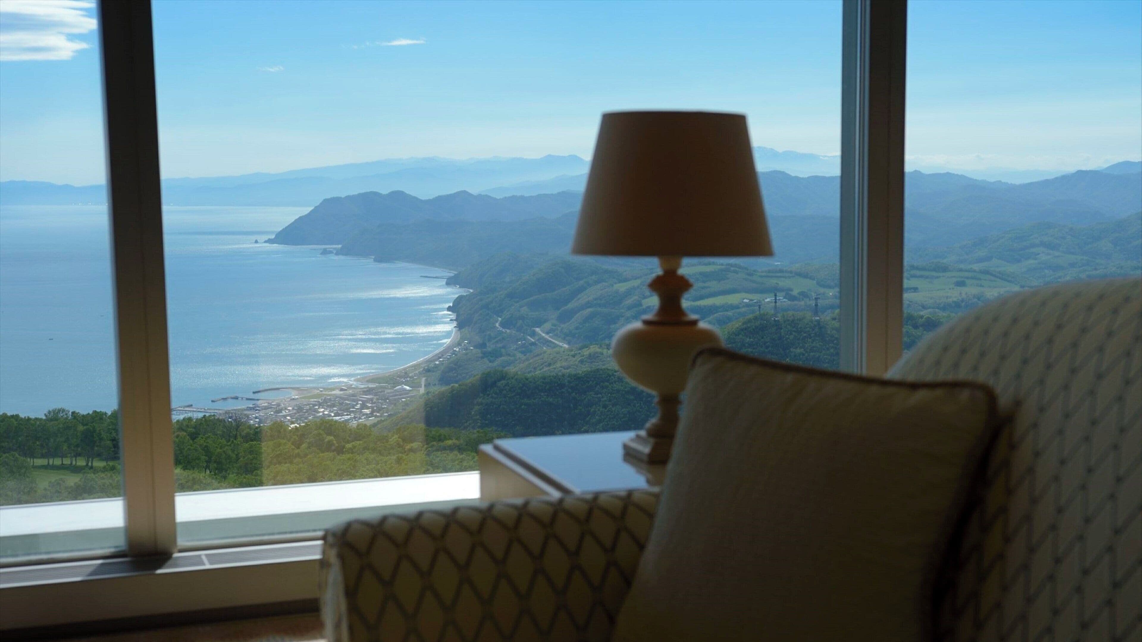A room with a large window offering a view of a coastal landscape, a lamp on a bedside table, and a patterned chair.