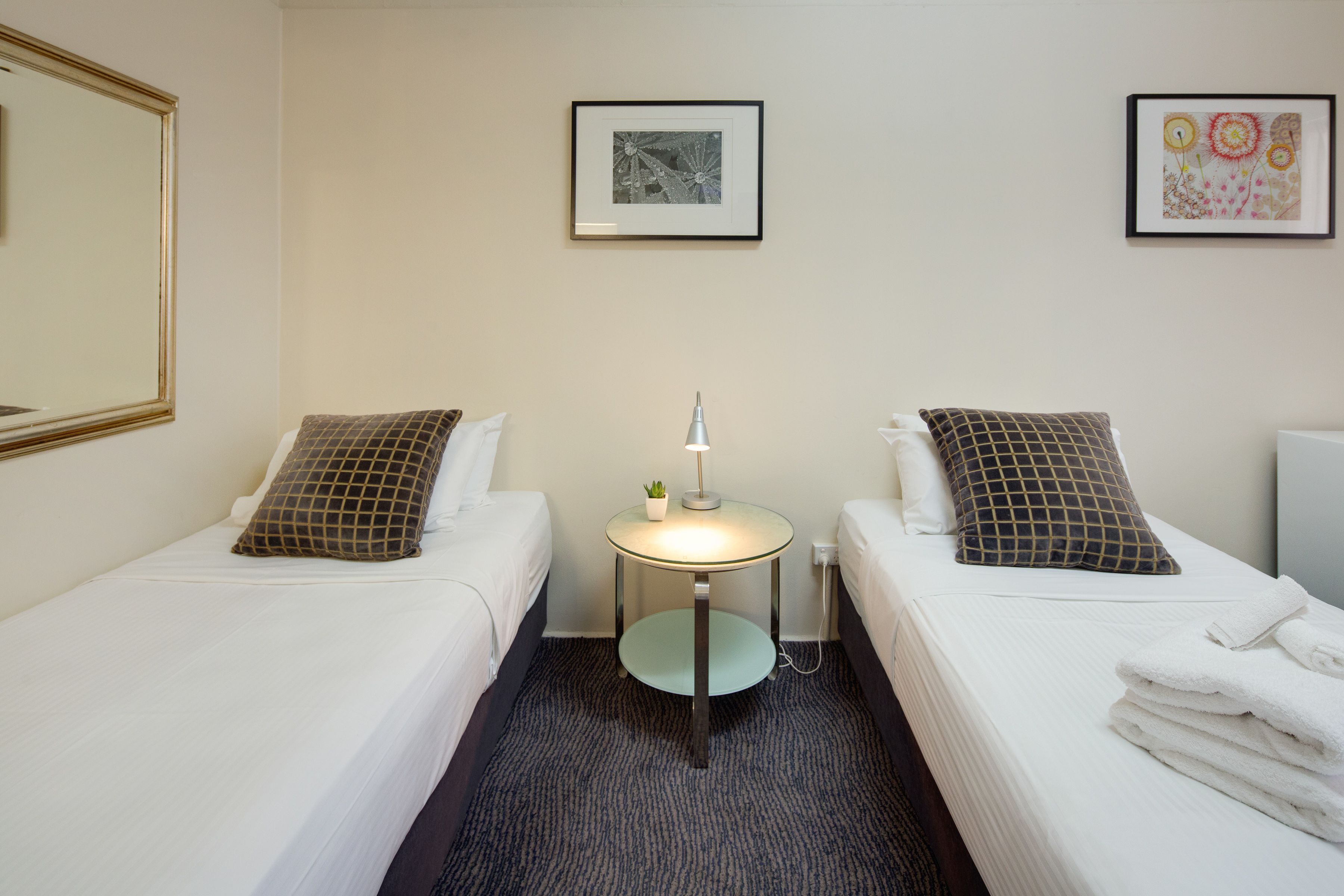A hotel room with two single beds, a small round table with a lamp, and two framed pictures on the wall.
