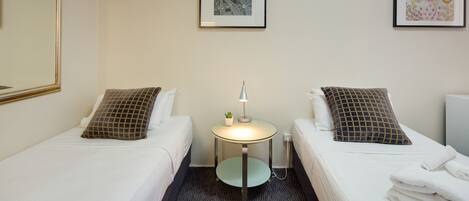 A hotel room with two single beds, a small round table with a lamp, and two framed pictures on the wall.