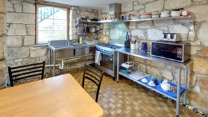 Eine Küche mit Steinwänden, einem Holztisch, Stühlen, einem Herd, einer Mikrowelle und Regalen mit Geschirr und Besteck.