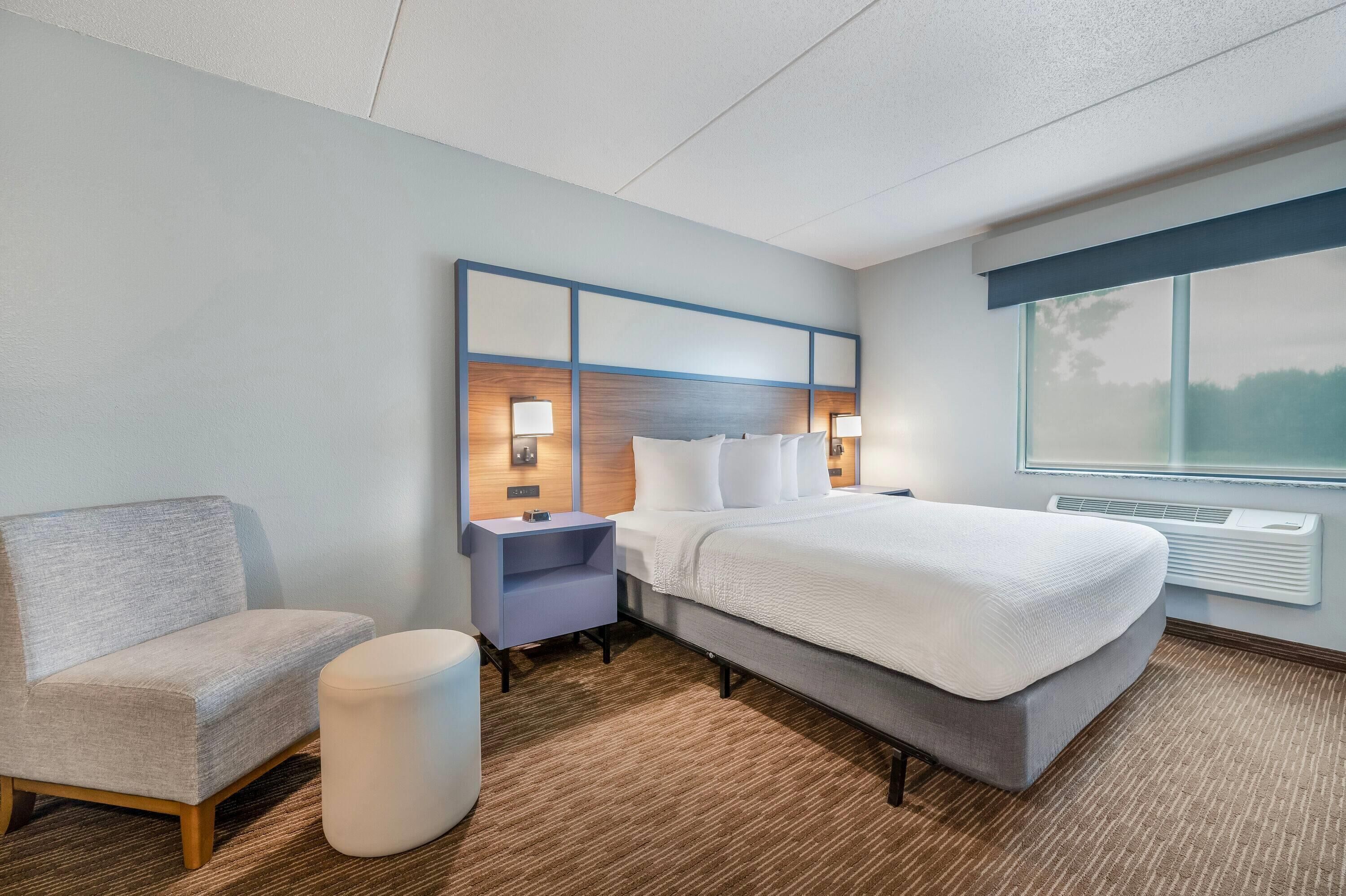 A hotel room with a bed, a nightstand, a chair, and a large window.