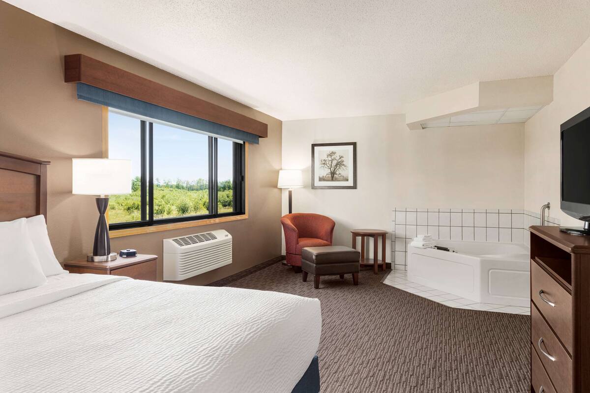 Habitación de hotel con cama, una silla, una mesita, televisor, bañera y ventana con vistas.