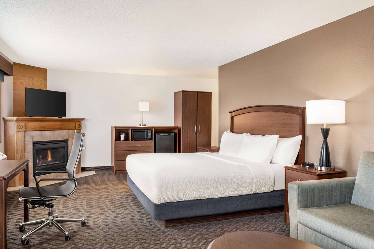 Habitación de hotel con una cama grande, un escritorio con silla, televisión, chimenea, armario y mesita de noche con lámpara.
