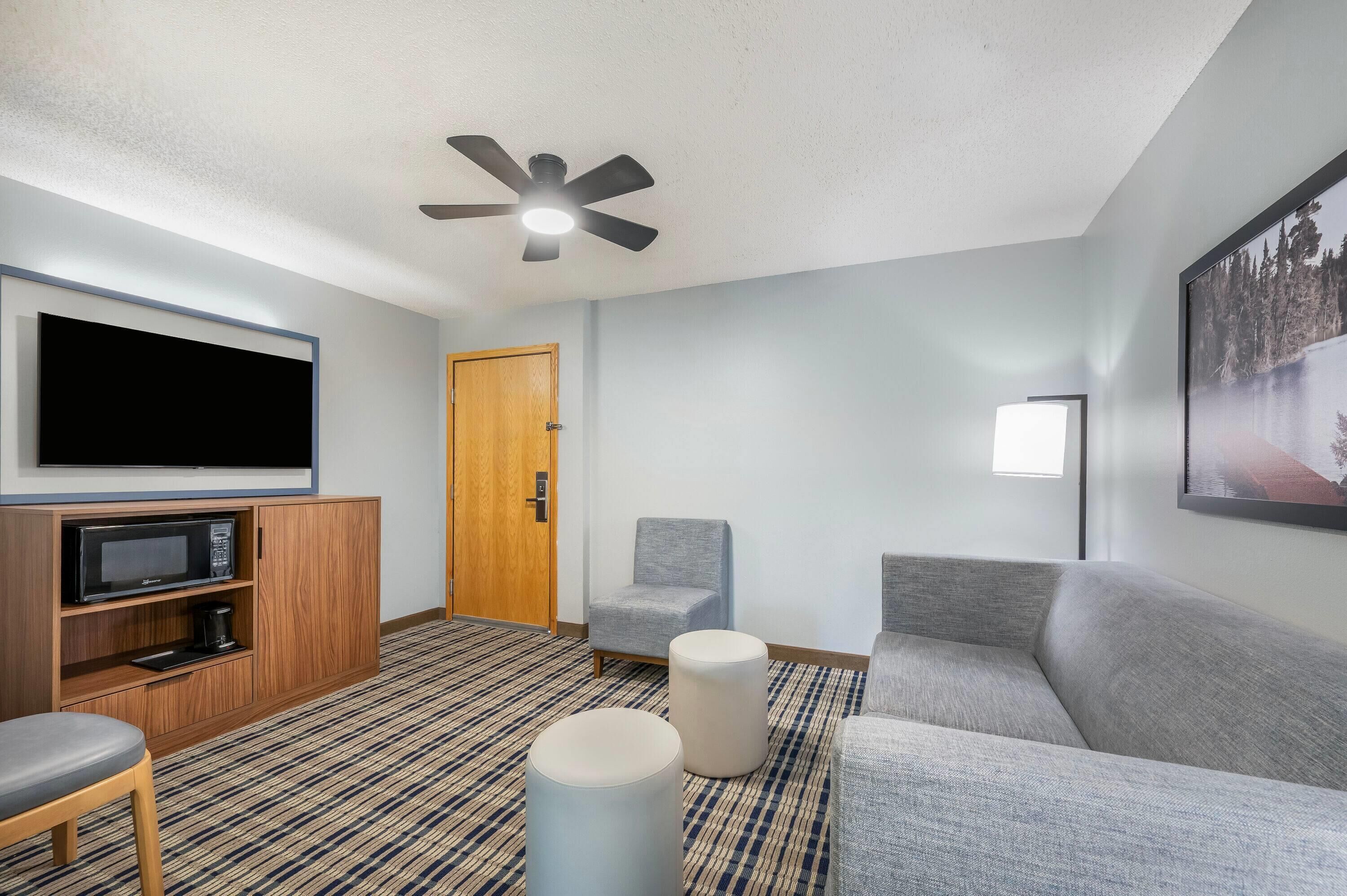 Habitación de hotel con televisor de pantalla plana, ventilador de techo, puerta de madera, sofá gris, sillón gris y alfombra estampada.