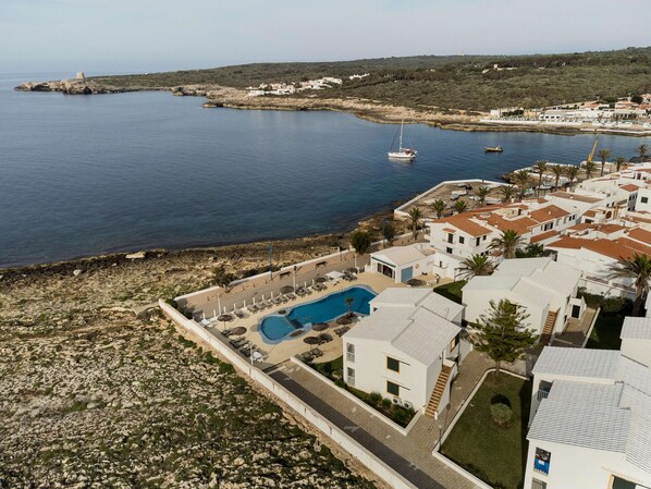 An aerial view of a coastal area with a swimming pool, white buildings, and a lighthouse in the distance. - Aluasun Far Menorca (Sant Lluis)