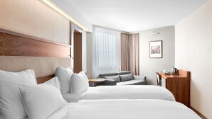 A hotel room with two beds, a sofa, a desk, and a large window with curtains. - Movenpick hotel Sarajevo (Sarajevo)