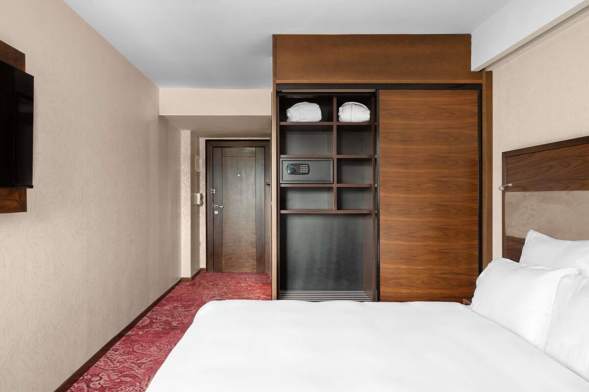 A hotel room with a large bed, a wooden wardrobe with shelves, a red carpet, and a door leading to another room.