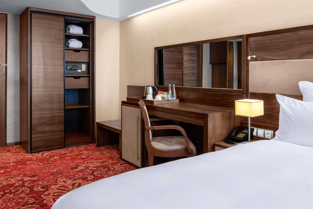 A hotel room with a bed, a desk with a chair, a minibar, a telephone, and a lamp.