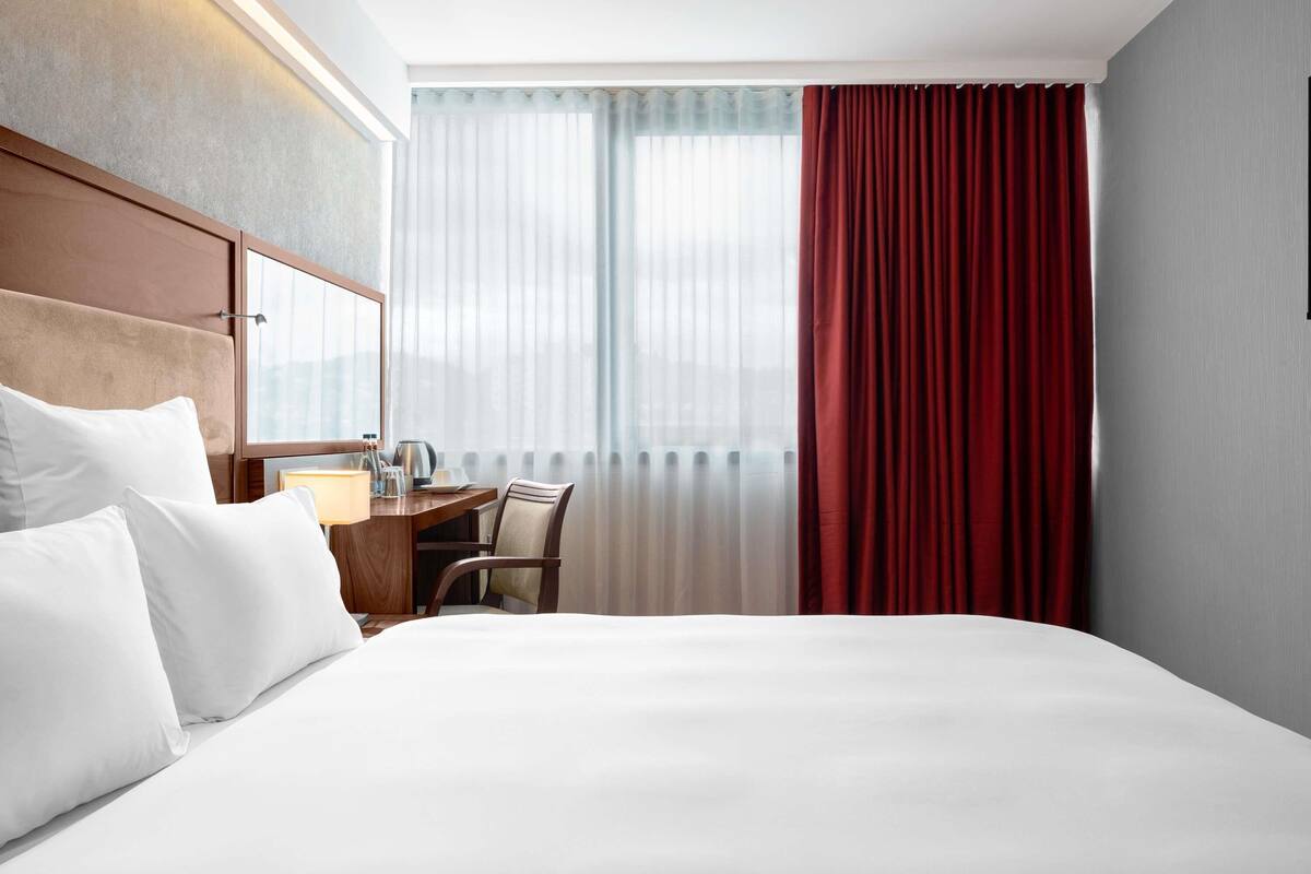 A hotel room with a large bed, a desk with a chair, a window with sheer curtains, and a red curtain.