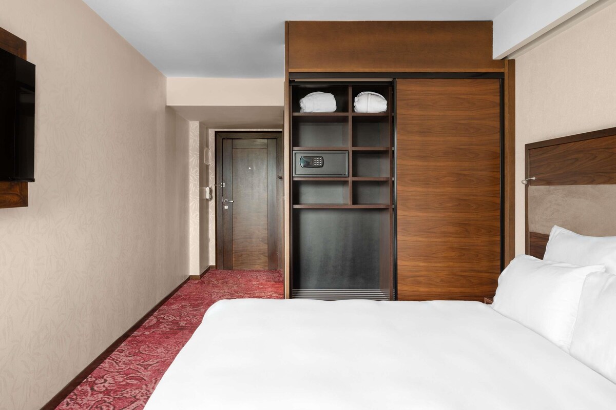 A hotel room with a large bed, a wooden wardrobe with shelves, a red carpet, and a door leading to another room.
