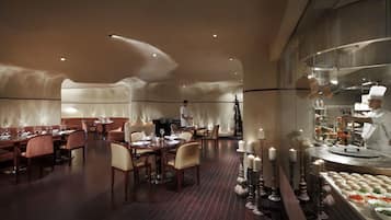 Een modern restaurant met een warme sfeer, met houten tafels en stoelen, kaarsdecoraties in het midden en een kok die in de achtergrond bereidt.