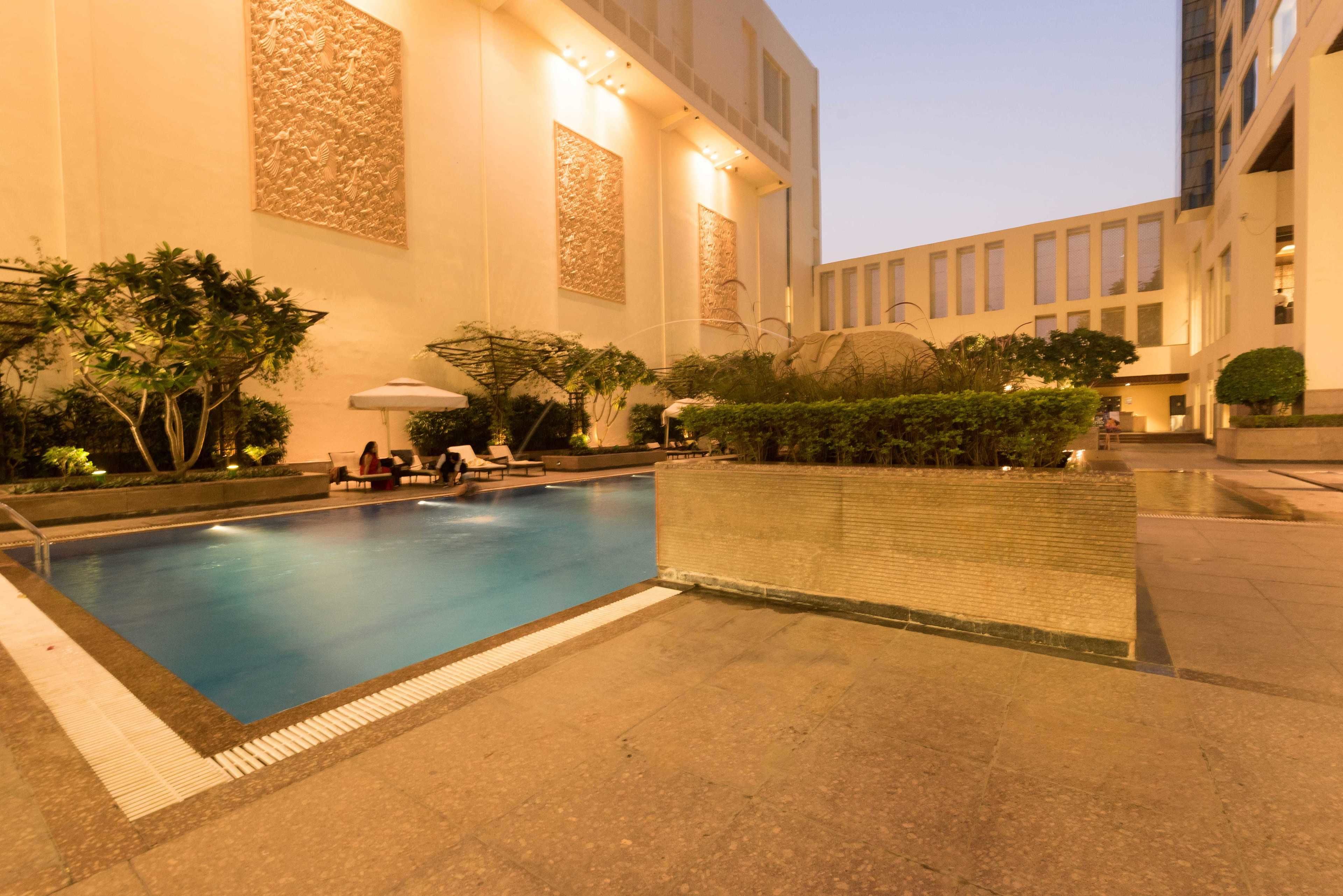 Piscina com espreguiçadeiras e guarda-sóis, cercada por plantas e um edifício com janelas amplas e painéis decorativos.