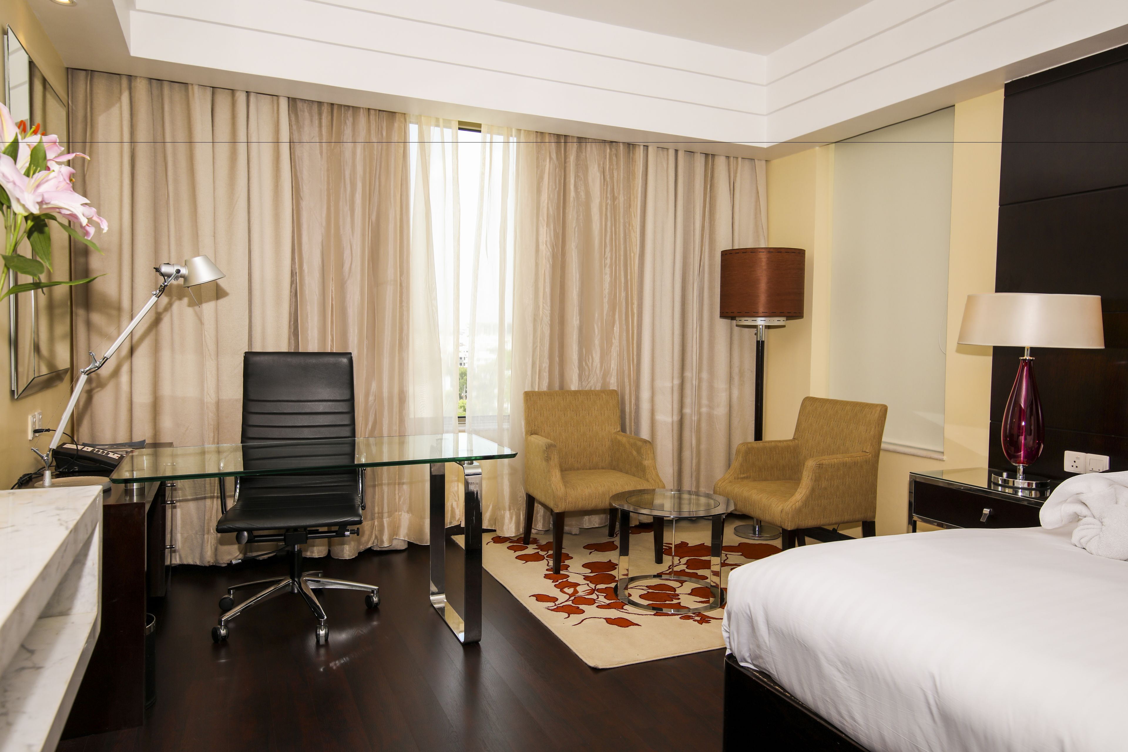 Habitación de hotel con una cama, un escritorio con silla, dos sillones, una mesita de vidrio, una lámpara y un ventanal con cortinas.