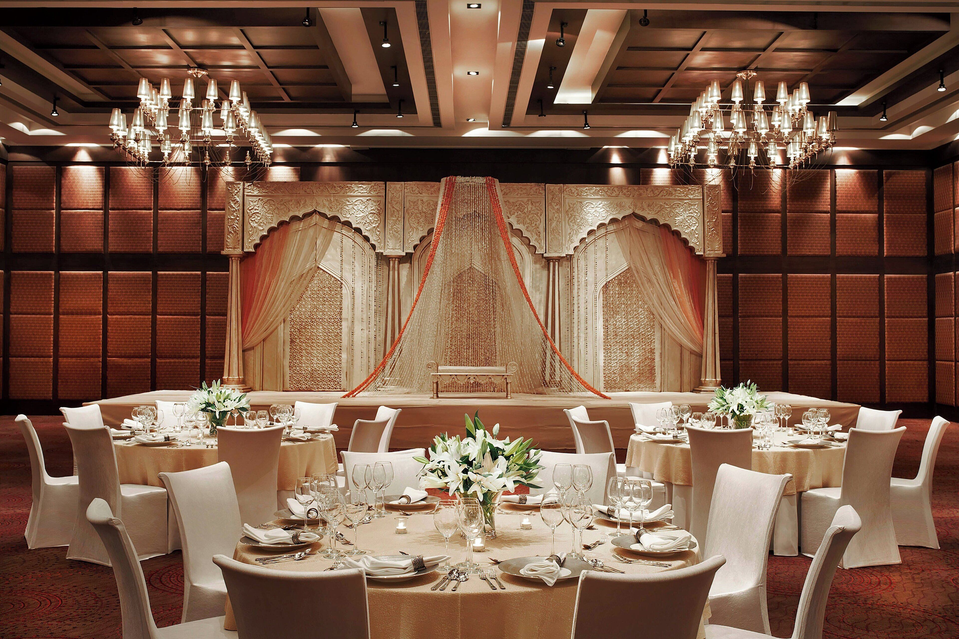 Un salón de banquetes con mesas redondas, sillas blancas, candelabros y un escenario con cortinas colgantes.