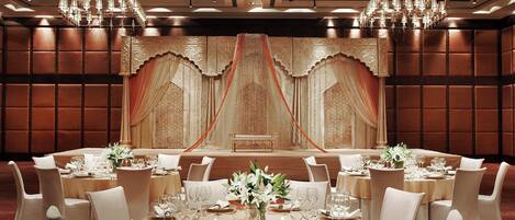 A banquet hall with round tables, white chairs, chandeliers, and a stage with draped curtains.