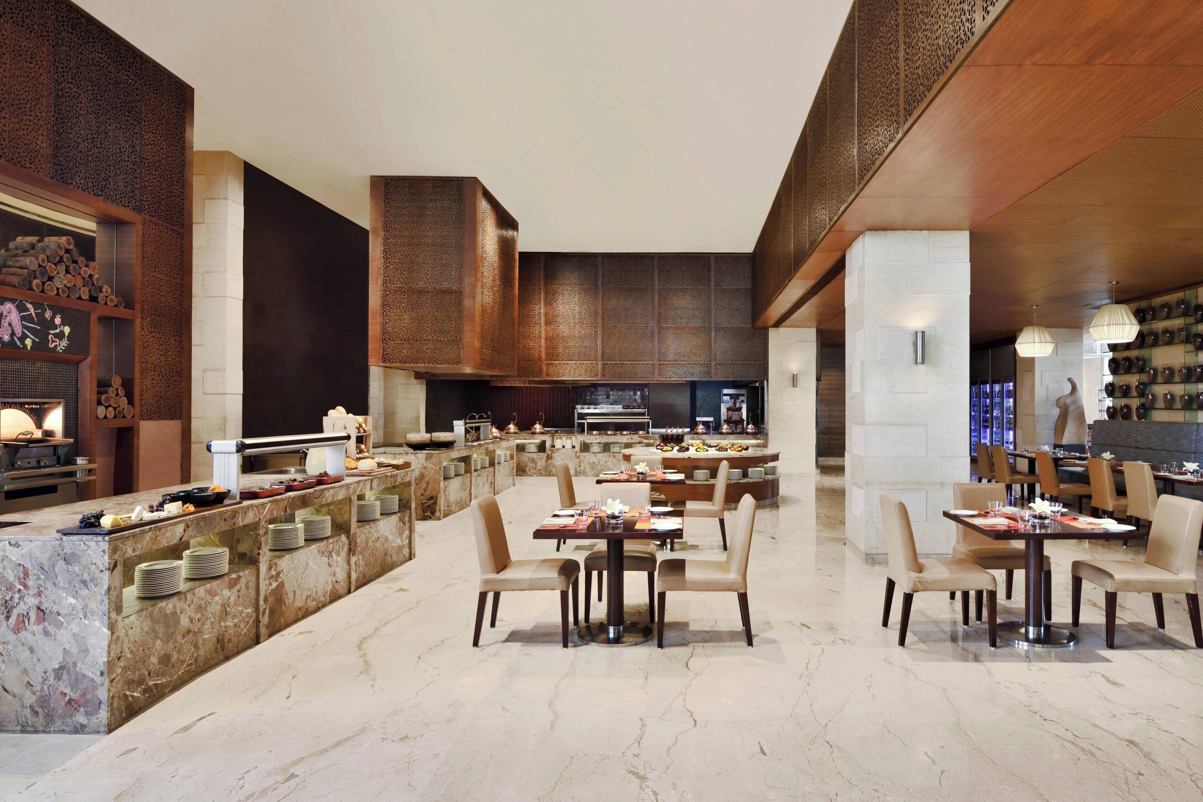 A spacious dining area with marble flooring, multiple tables and chairs, a buffet counter, and a modern kitchen in the background.