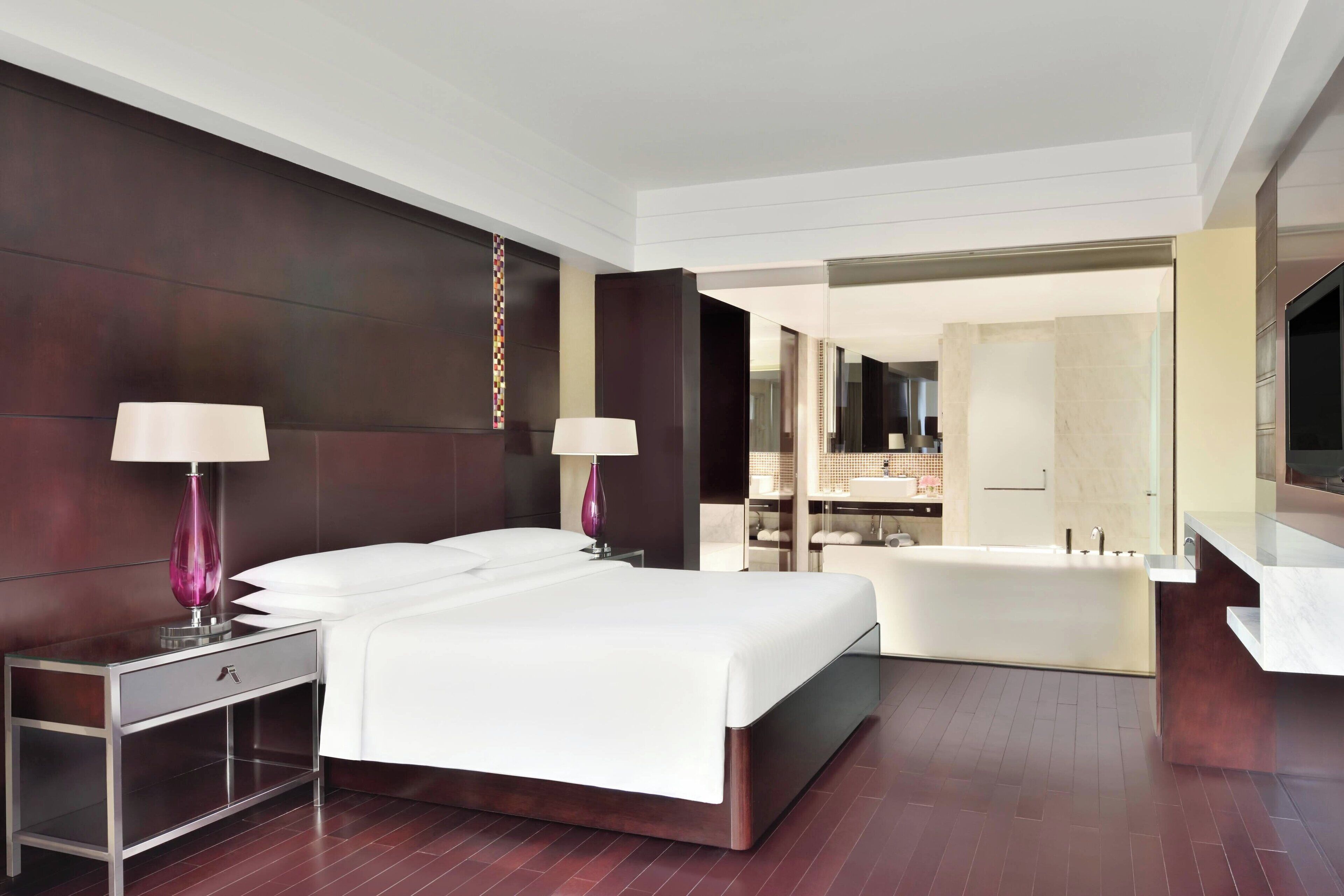 A modern hotel room with a large bed, two bedside tables with lamps, a built-in bathtub, and a large mirror.